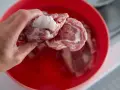 Ręka wkłada surowe mięso do czerwonej miski z wodą, aby szybko rozmrozić mięso.