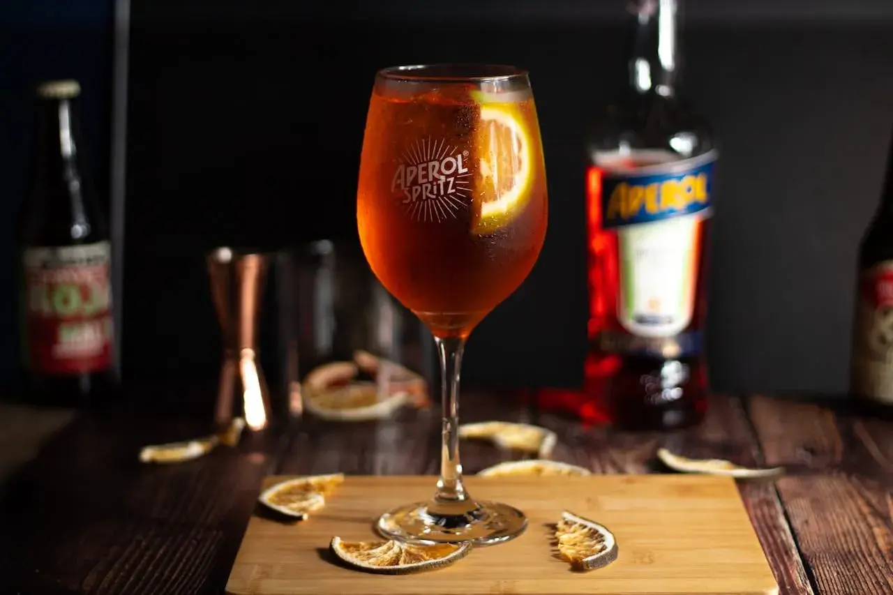 Orzeźwiający aperol spritz z plasterkiem pomarańczy, otoczony suszonymi owocami i butelką Aperolu. Idealny na letnie popołudnie.
