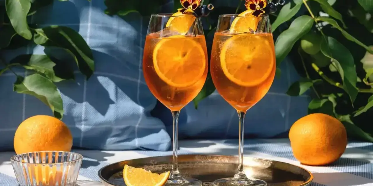 Dwa orzeźwiające drinki z aperolem i sokiem pomarańczowym, ozdobione plastrami pomarańczy i kwiatami, gotowe na letnie popołudnie.