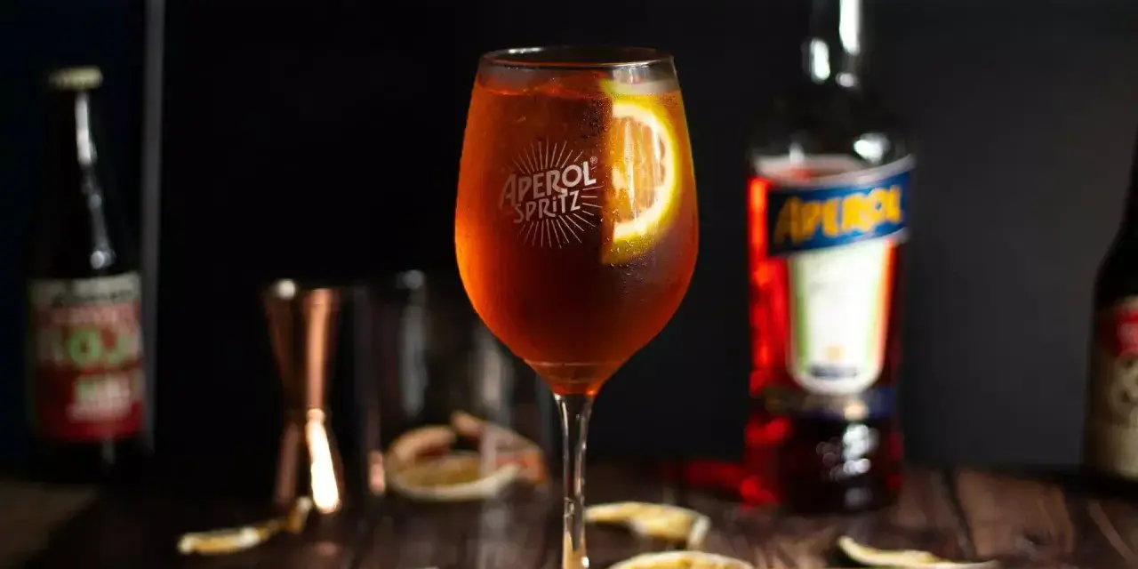 Orzeźwiający drink z aperolem, ozdobiony plasterkiem pomarańczy. Obok butelka Aperol Spritz i inne akcesoria barmańskie.