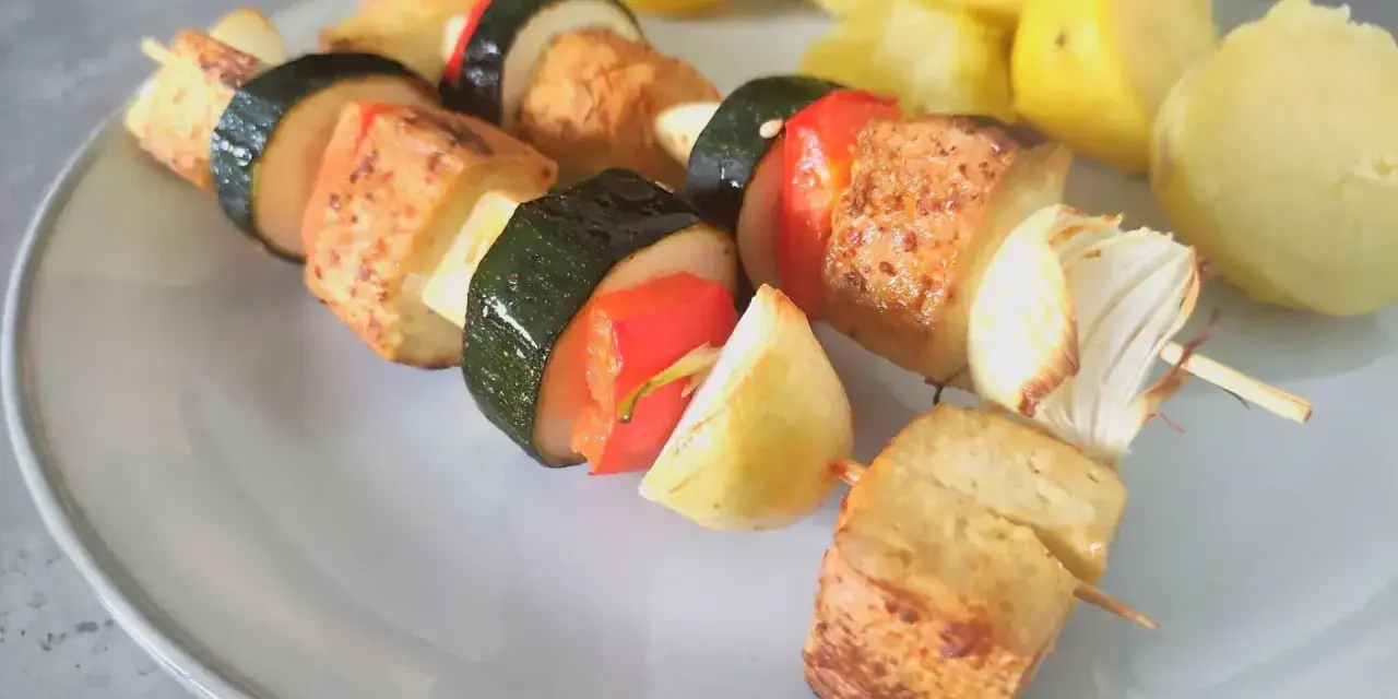 Szaszłyki z tofu, cukinii, papryki i cebuli na talerzu, obok gotowane ziemniaki.