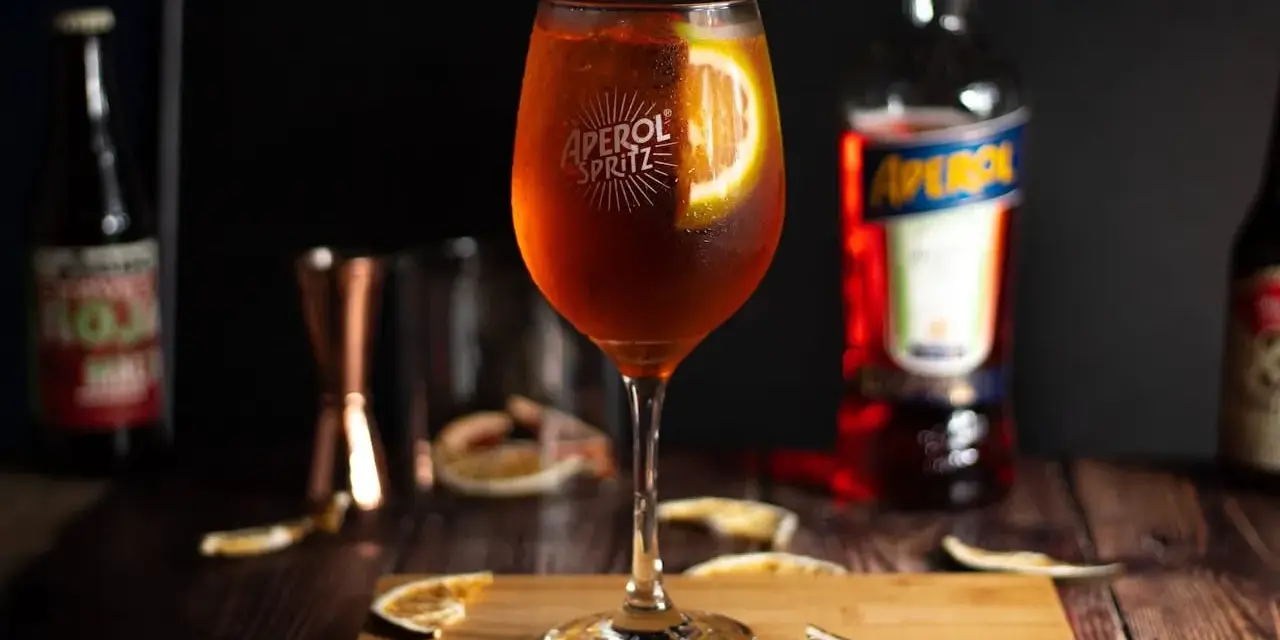 Orzeźwiający aperol spritz z plasterkiem pomarańczy, otoczony suszonymi owocami i butelką Aperolu. Idealny na letnie popołudnie.