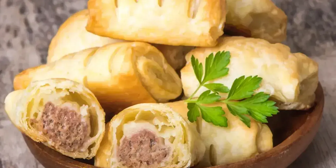 Stos pieczonego ciasta francuskiego z kiełbasą w środku, ozdobione gałązką pietruszki. Idealne na szybką przekąskę.