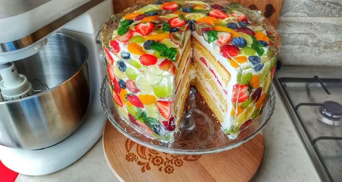 Kolorowy tort z owocami, z kawałkiem wyciętym, prezentuje się apetycznie. Obok stoi mikser.
