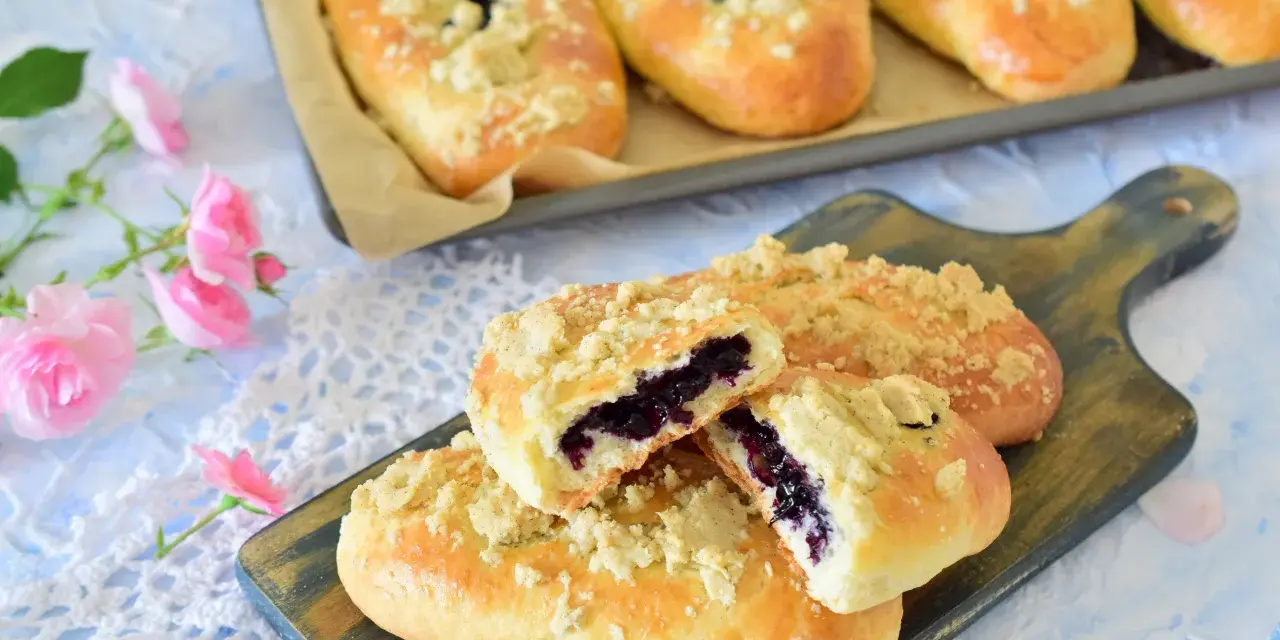 Pyszne jagodzianki z kruszonką, jedna przekrojona, ukazująca soczyste jagody. Obok różowe róże.