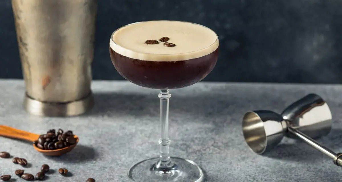 Elegancki drink z kawą, ozdobiony ziarnami kawy, obok shakera i miarki barmańskiej. Idealny na pobudzenie.