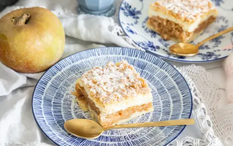 Pyszny jabłecznik z budyniem, posypany cukrem pudrem, czeka na talerzu. Obok leży jabłko.