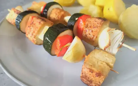 Szaszłyki z tofu, cukinii, papryki i cebuli na talerzu, obok gotowane ziemniaki.