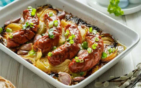 Pieczona biała kiełbasa surowa z cebulą i czosnkiem w naczyniu żaroodpornym, posypana szczypiorkiem.