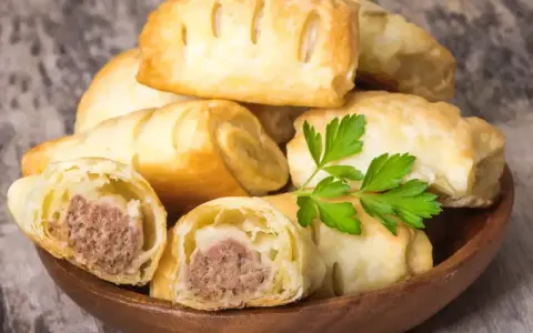 Stos pieczonego ciasta francuskiego z kiełbasą w środku, ozdobione gałązką pietruszki. Idealne na szybką przekąskę.