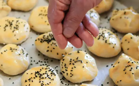 Ręka posypuje czarnuszką gotowe do pieczenia bułki z serka wiejskiego.