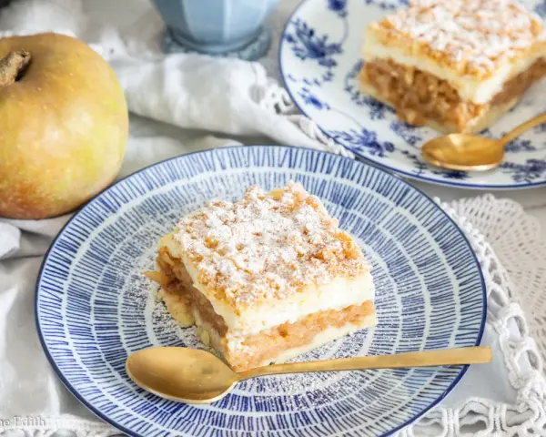 Pyszny jabłecznik z budyniem, posypany cukrem pudrem, czeka na talerzu. Obok leży jabłko.