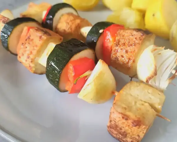 Szaszłyki z tofu, cukinii, papryki i cebuli na talerzu, obok gotowane ziemniaki.