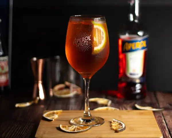 Orzeźwiający aperol spritz z plasterkiem pomarańczy, otoczony suszonymi owocami i butelką Aperolu. Idealny na letnie popołudnie.