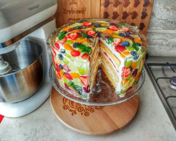 Kolorowy tort z owocami, z kawałkiem wyciętym, prezentuje się apetycznie. Obok stoi mikser.