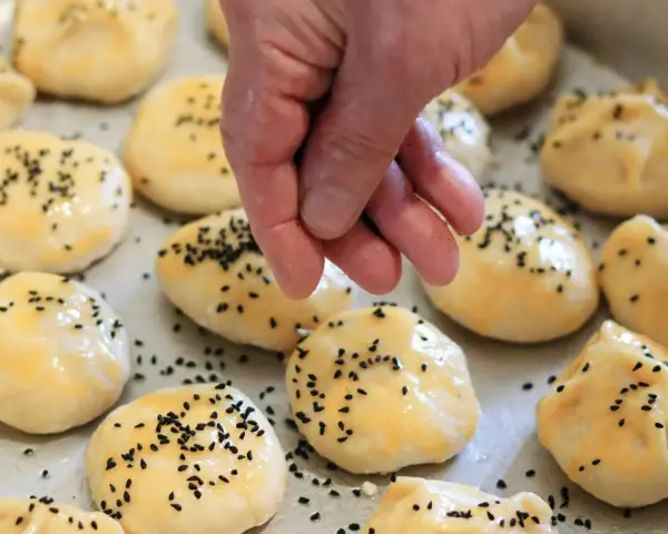 Ręka posypuje czarnuszką gotowe do pieczenia bułki z serka wiejskiego.