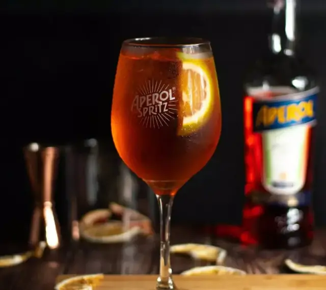 Orzeźwiający drink z aperolem, ozdobiony plasterkiem pomarańczy. Obok butelka Aperol Spritz i inne akcesoria barmańskie.