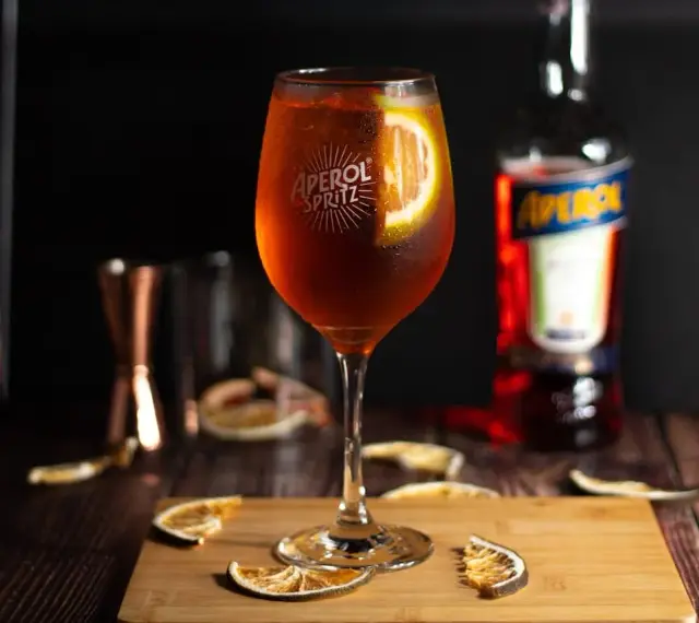 Orzeźwiający aperol spritz z plasterkiem pomarańczy, otoczony suszonymi owocami i butelką Aperolu. Idealny na letnie popołudnie.