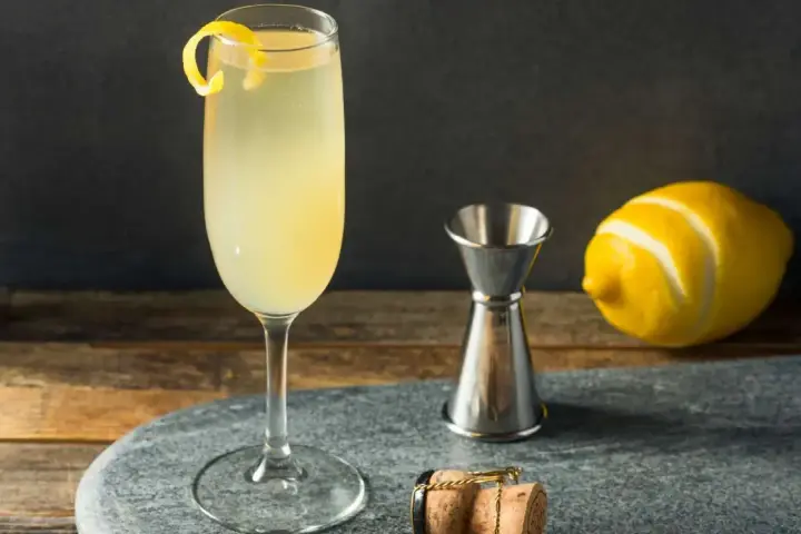 Elegancki drink z szampanem, ozdobiony skórką cytryny, obok kieliszka stoi miarka barmańska, cytryna i korek od szampana.