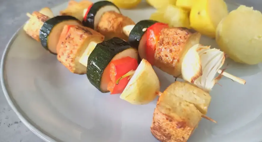 Szaszłyki z tofu, cukinii, papryki i cebuli na talerzu, obok gotowane ziemniaki.