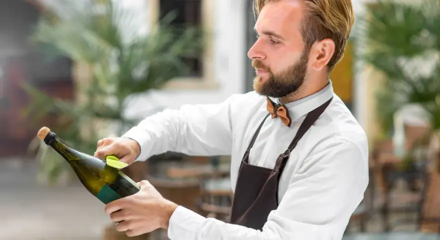 Barman w białej koszuli i brązowym fartuchu pokazuje, jak otwierać szampana, trzymając butelkę i wyciągając korek.