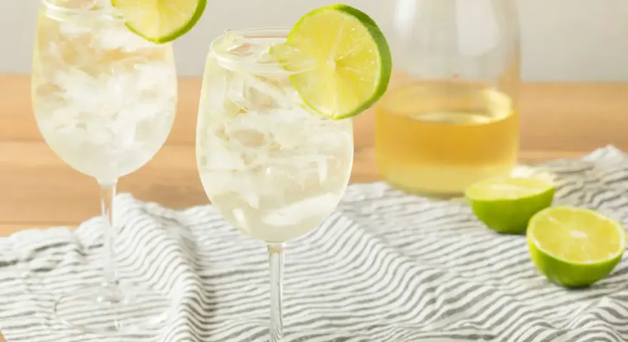Orzeźwiający szprycer co to? Dwa kieliszki białego wina z lodem i plasterkiem limonki, obok butelka wina i połówki limonki.