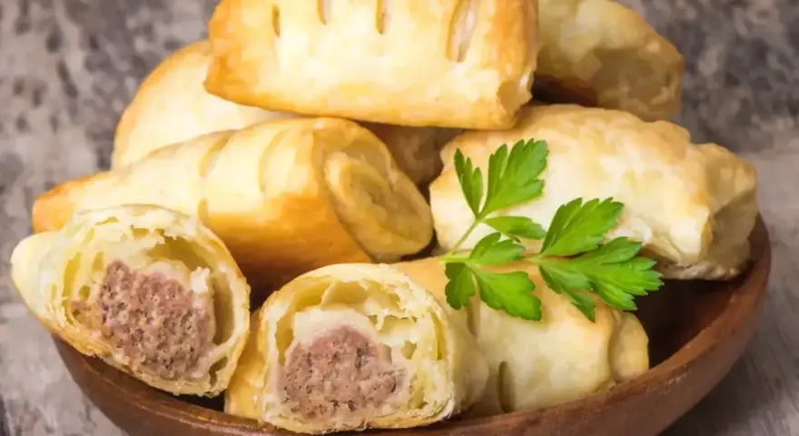 Stos pieczonego ciasta francuskiego z kiełbasą w środku, ozdobione gałązką pietruszki. Idealne na szybką przekąskę.