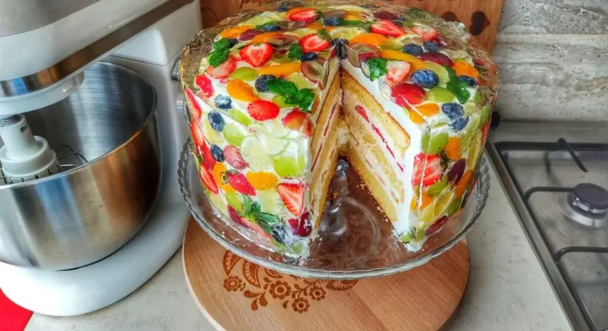 Kolorowy tort z owocami, z kawałkiem wyciętym, prezentuje się apetycznie. Obok stoi mikser.