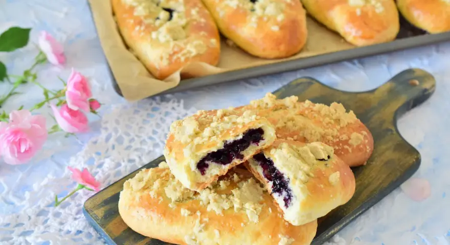 Pyszne jagodzianki z kruszonką, jedna przekrojona, ukazująca soczyste jagody. Obok różowe róże.