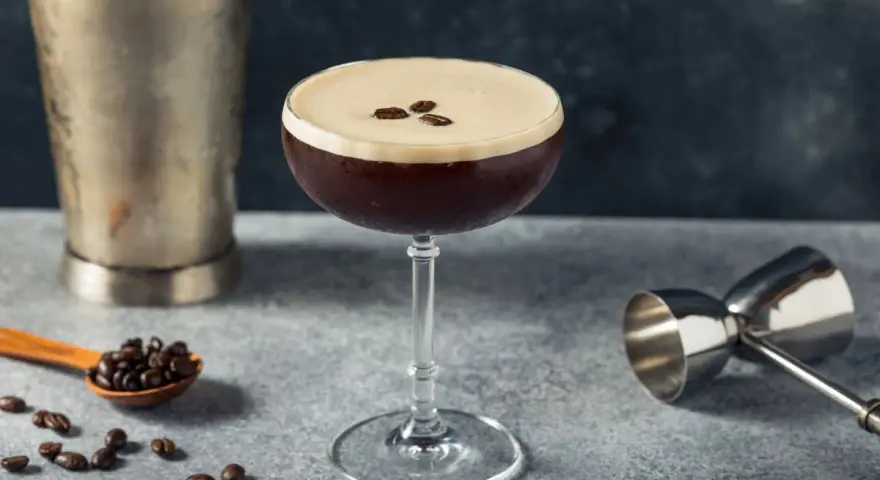 Elegancki drink z kawą, ozdobiony ziarnami kawy, obok shakera i miarki barmańskiej. Idealny na pobudzenie.
