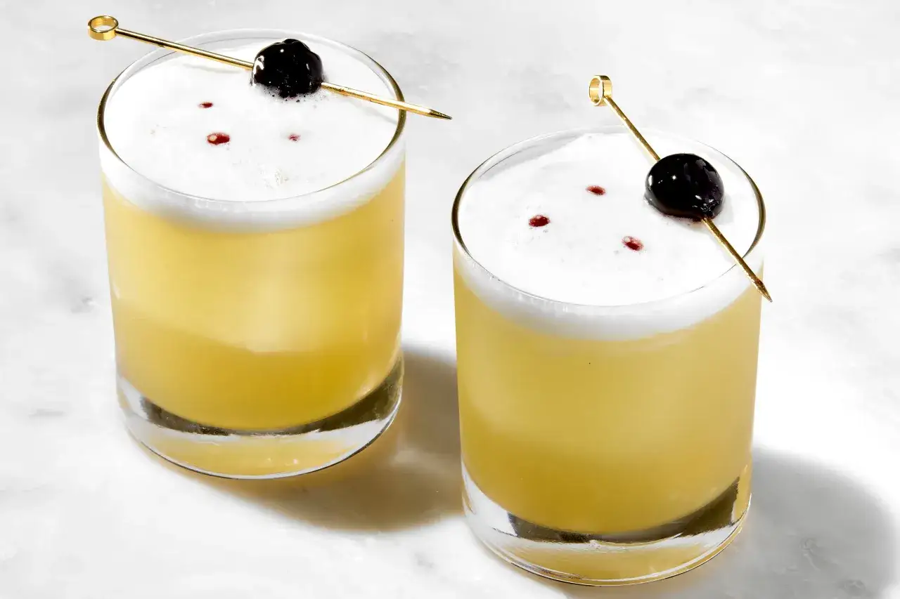 Dwa drinki whisky sour z pianką, wisienką i złotym patyczkiem na marmurowym blacie.