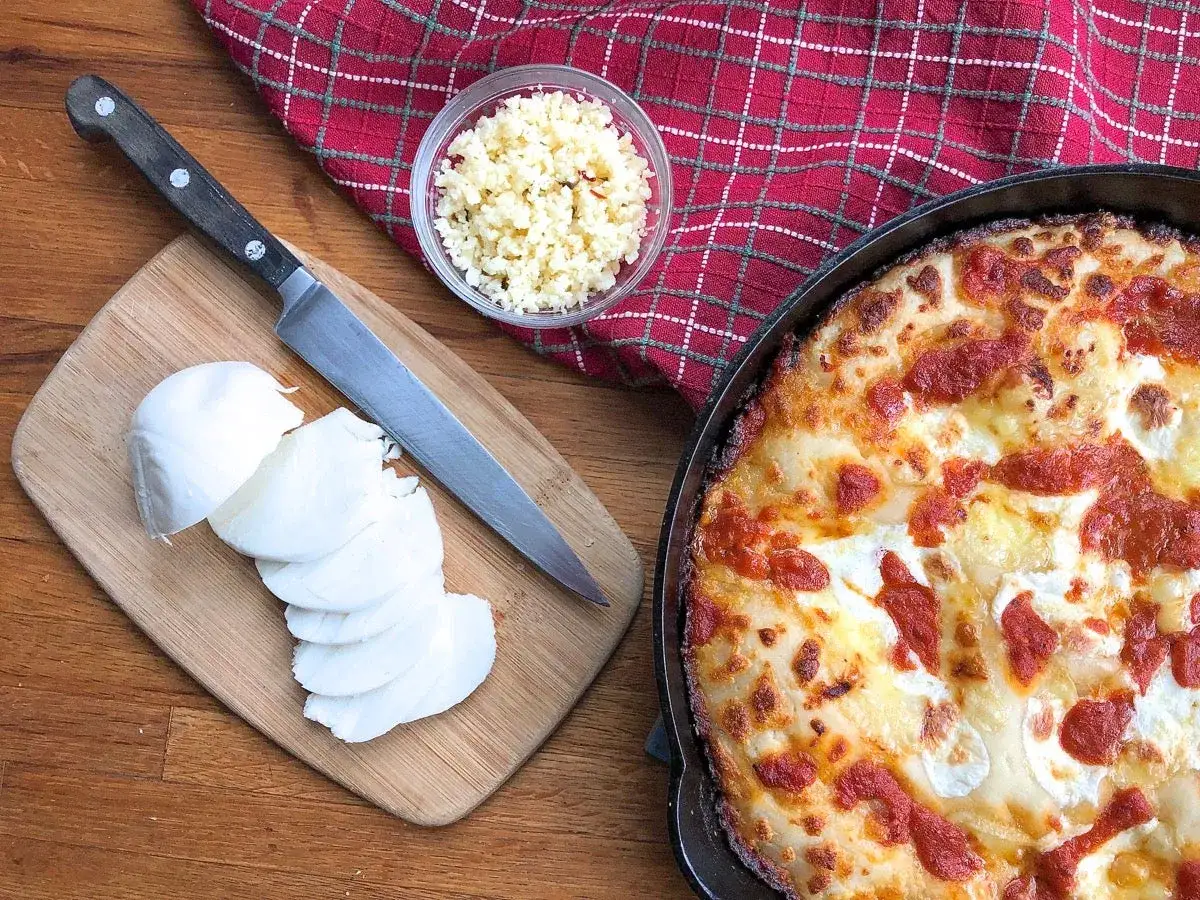 Składniki na pyszną pizzę: świeży ser, nóż i miseczka z tartym serem. Gotowa pizza w tle.