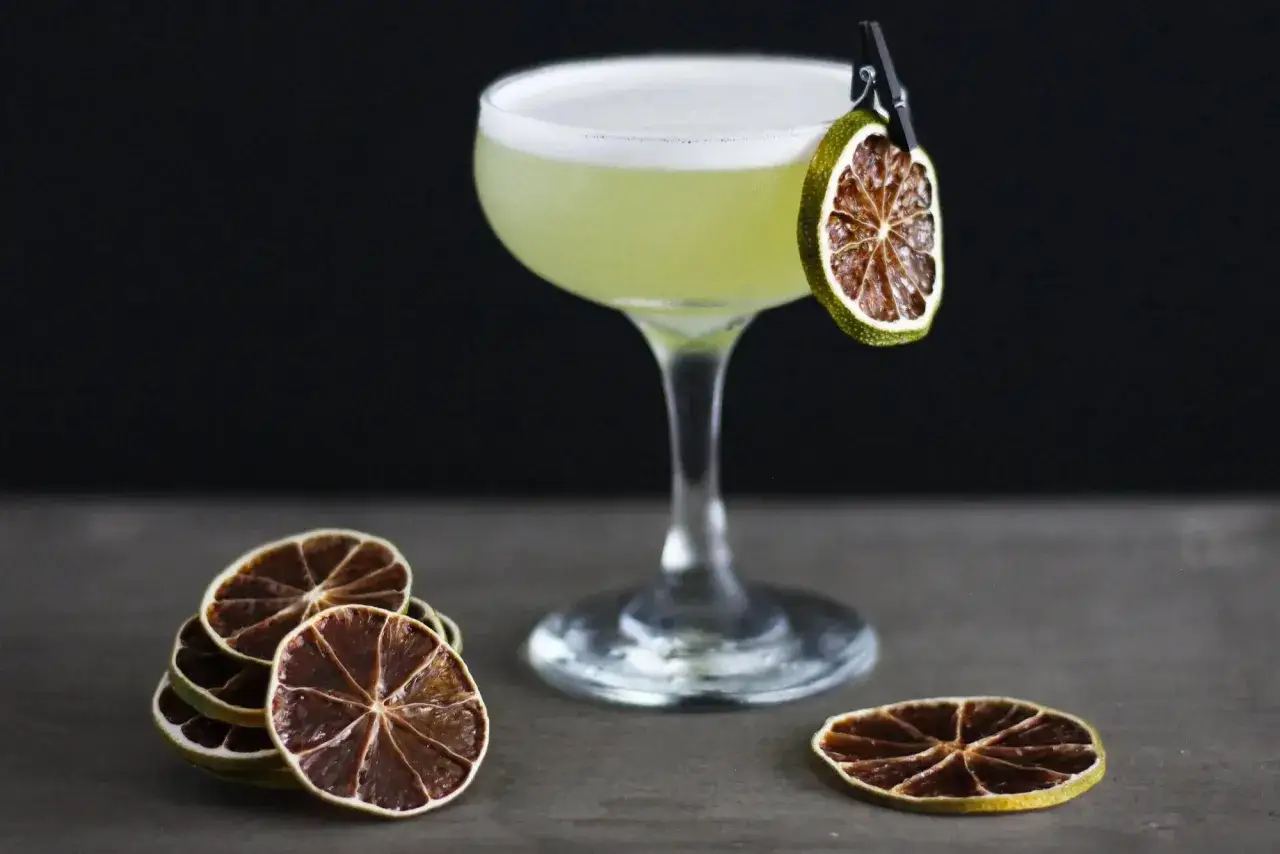 Zielony, orzeźwiający drink z pianką, ozdobiony suszoną limonką na klipsie. Obok stosik plasterków cytrusów. Idealny na letni wieczór, ostatnie słowo cocktail.