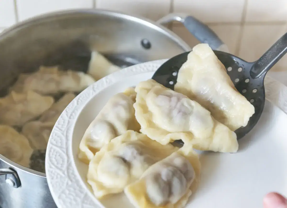 Gotowanie pierogów: wyjmowanie z garnka ugotowanych, parujących pierogów na biały talerz.