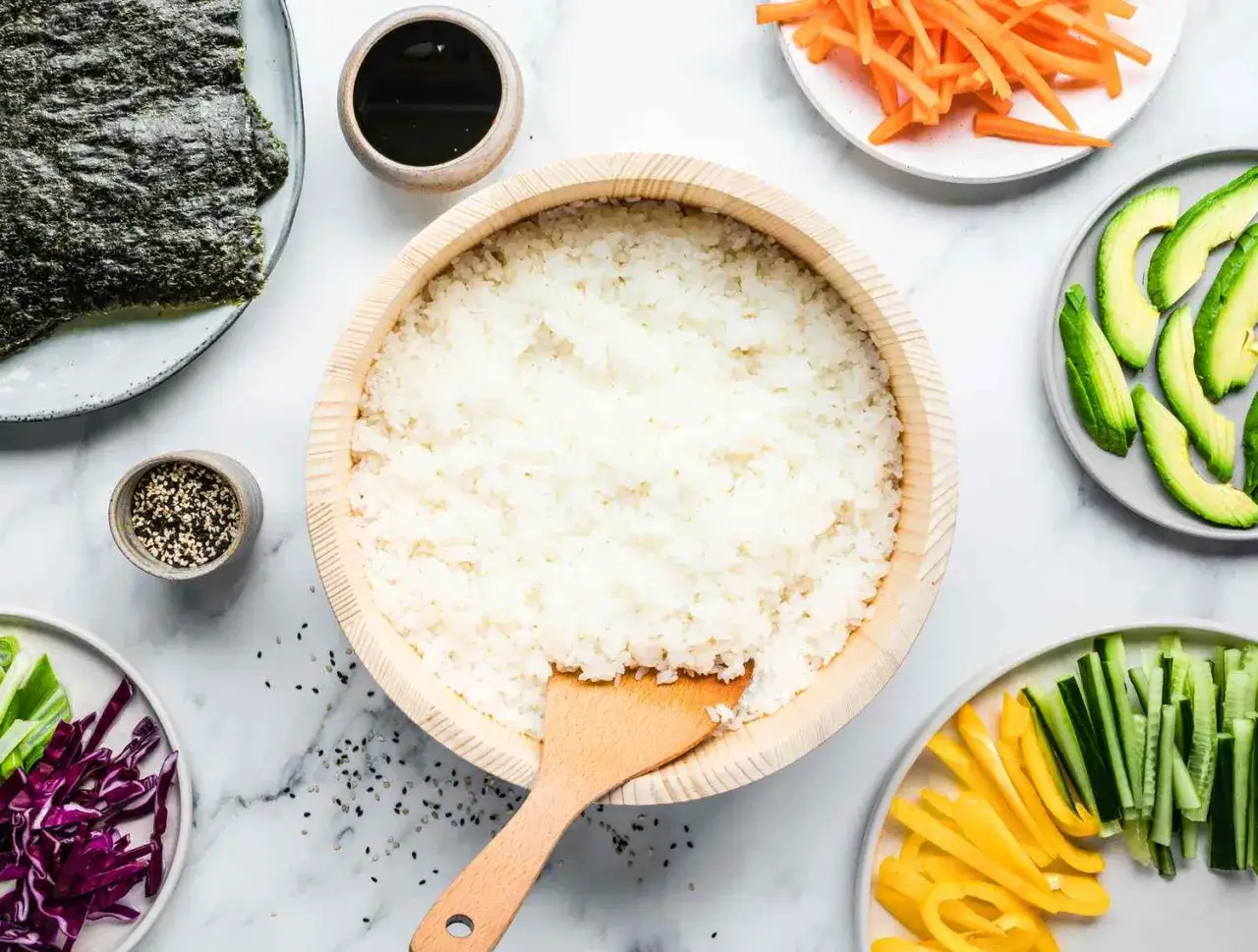 Idealny ryż do sushi w drewnianej misce, otoczony nori, awokado, marchewką, ogórkiem, papryką i sezamem.