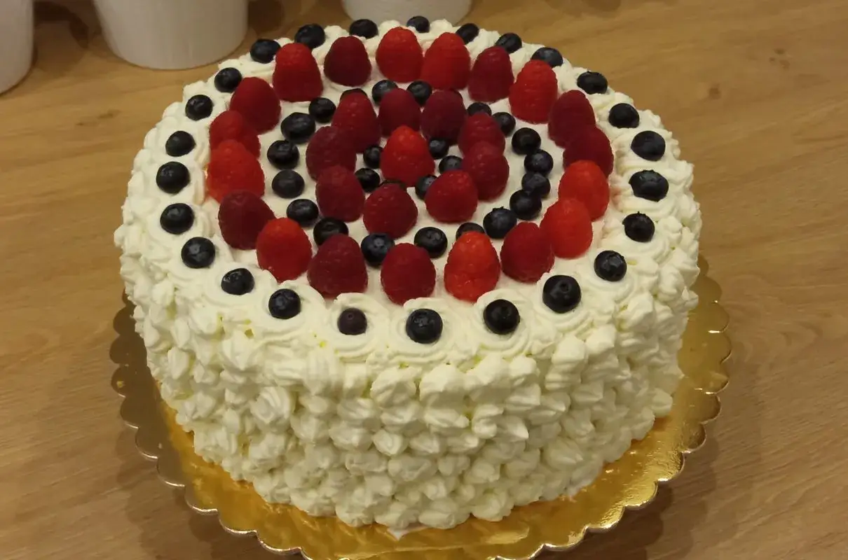 Pyszny tort bezglutenowy ozdobiony bitą śmietaną, malinami i borówkami. Idealny na każdą okazję, a przepis jest prosty!