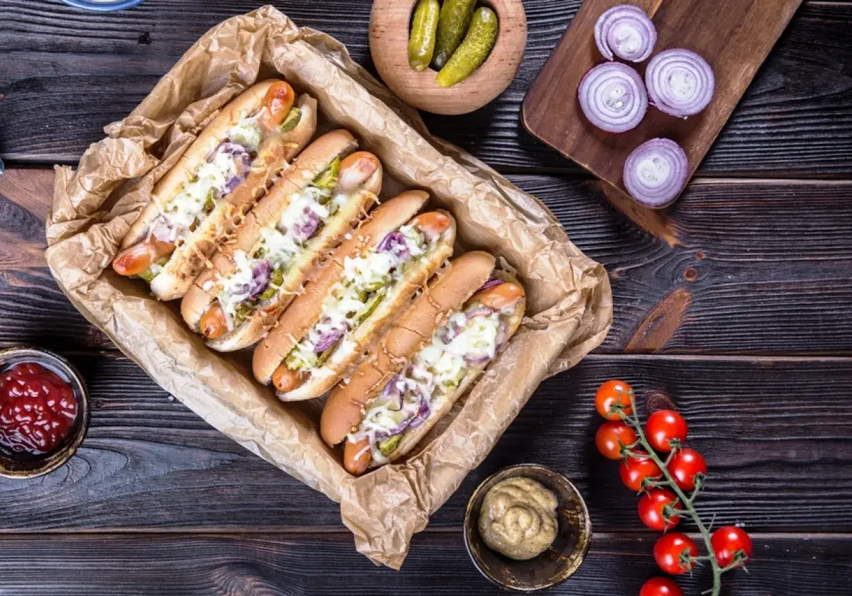 Pięć apetycznych hot dog&oacute;w domowych z serem, og&oacute;rkiem i czerwoną cebulą, podanych na brązowym papierze. Obok ketchup, musztarda i pomidorki.