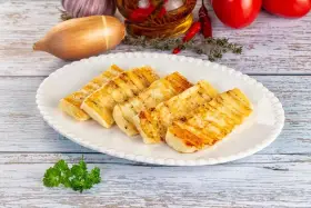 Grillowany ser halloumi na białym talerzu, z dodatkami jak czosnek, cebula i pomidory w tle.
