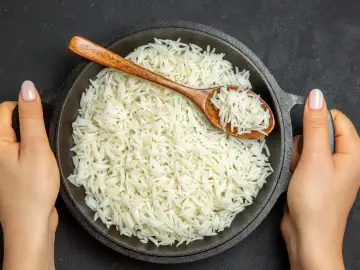 Dwie ręce trzymają okrągłą, czarną patelnię pełną puszystego ryżu basmati. Drewniana łyżka zanurzona w ryżu.